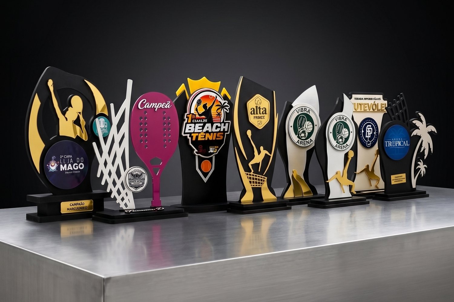 Nove modelos de troféus personalizados da Podium Premiações lado a lado sobre uma bancada metálica, com peças de beach tennis, futevôlei e futebol